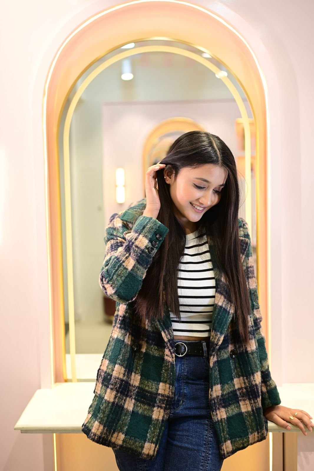 Woman in a green check jacket with long hair, in front of an arched gold-framed mirror