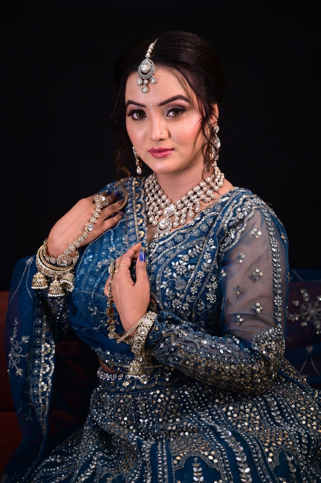 Woman in a blue embroidered outfit with a layered necklace and maang tikka against a dark backdrop