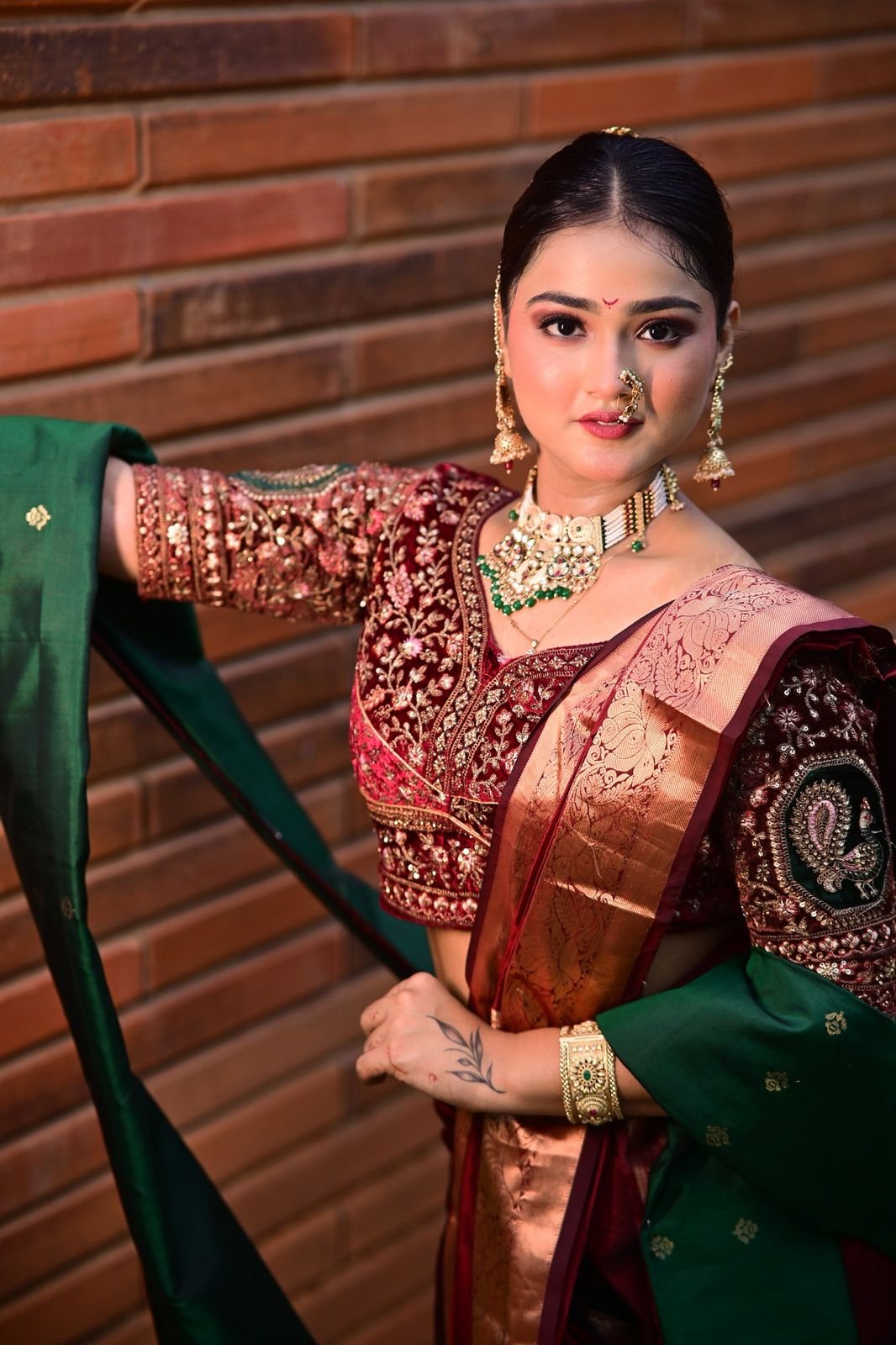 Bride in a maroon blouse with a green dupatta and nath, leaning against a brick wall