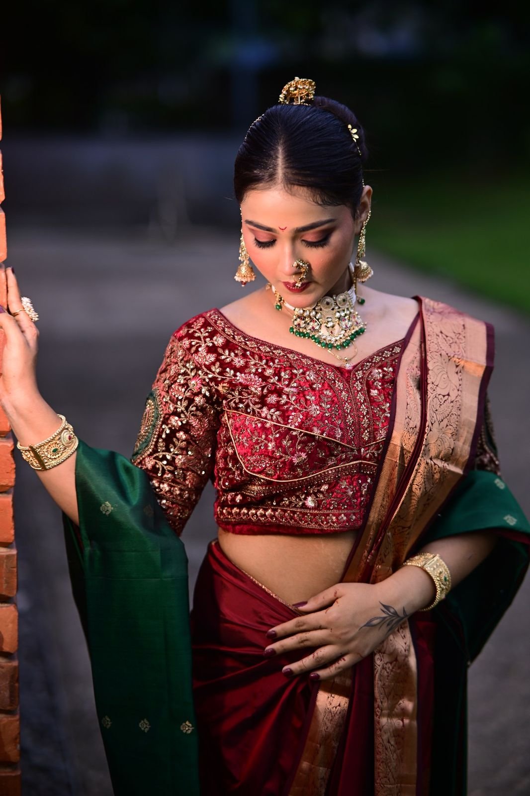 Bride in a maroon blouse and saree, standing against a brick wall