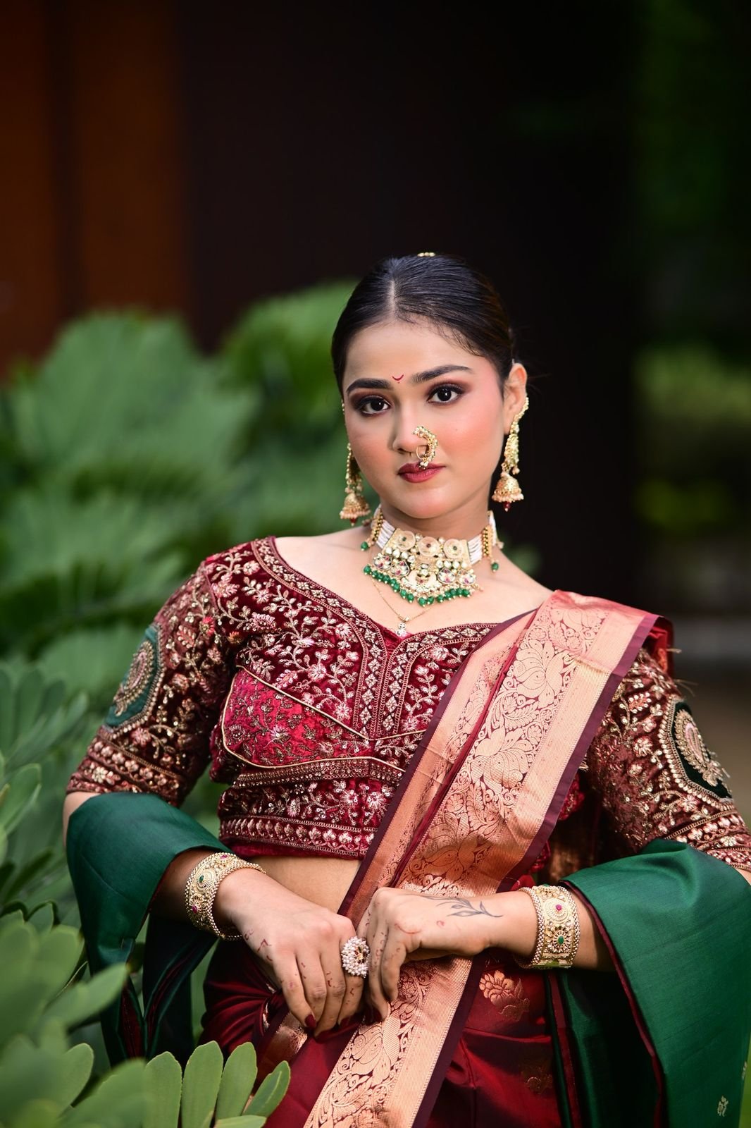 Bride in a maroon blouse with a green dupatta and kundan necklace, posed beside green leaves