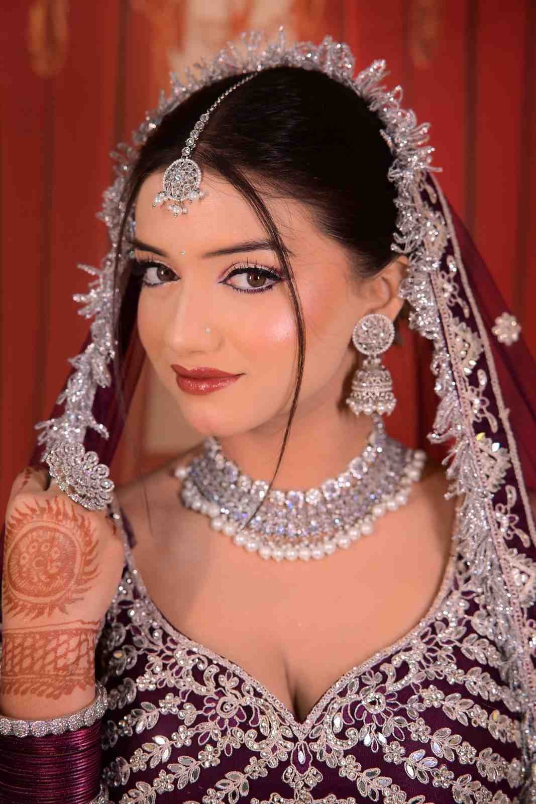 Bride in a maroon lehenga with a silver-white veil, jewellery and mehndi against a red curtain