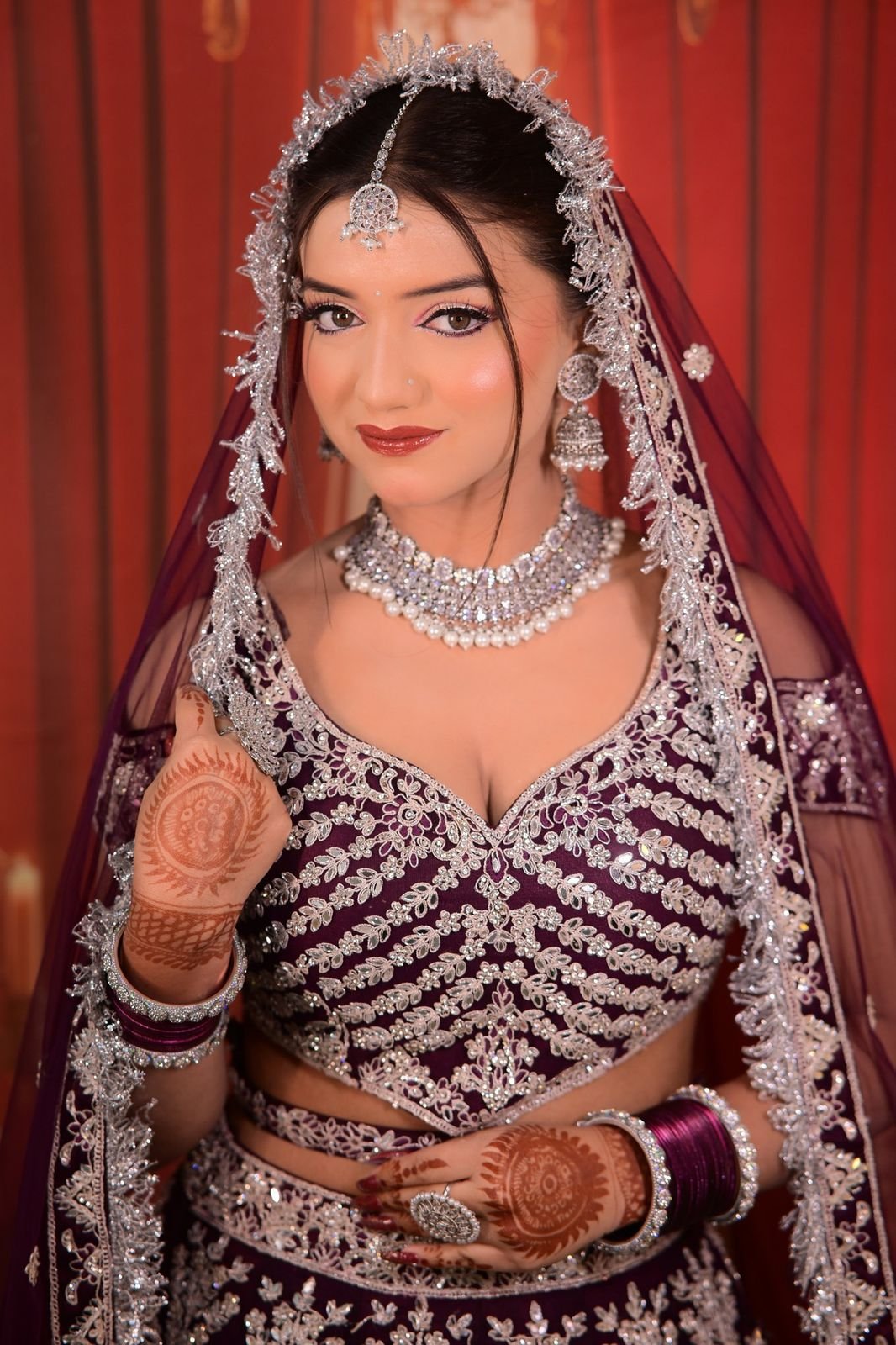 Bride in a plum embroidered lehenga with a silver-trimmed veil, mehndi and a red bangle set against a red curtain
