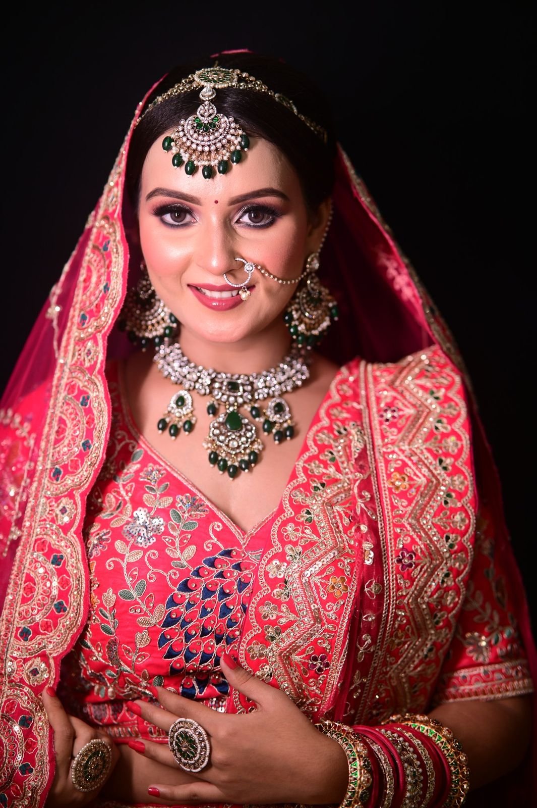 Bride in a red embroidered lehenga and veil with a green kundan set, photographed against a dark backdrop