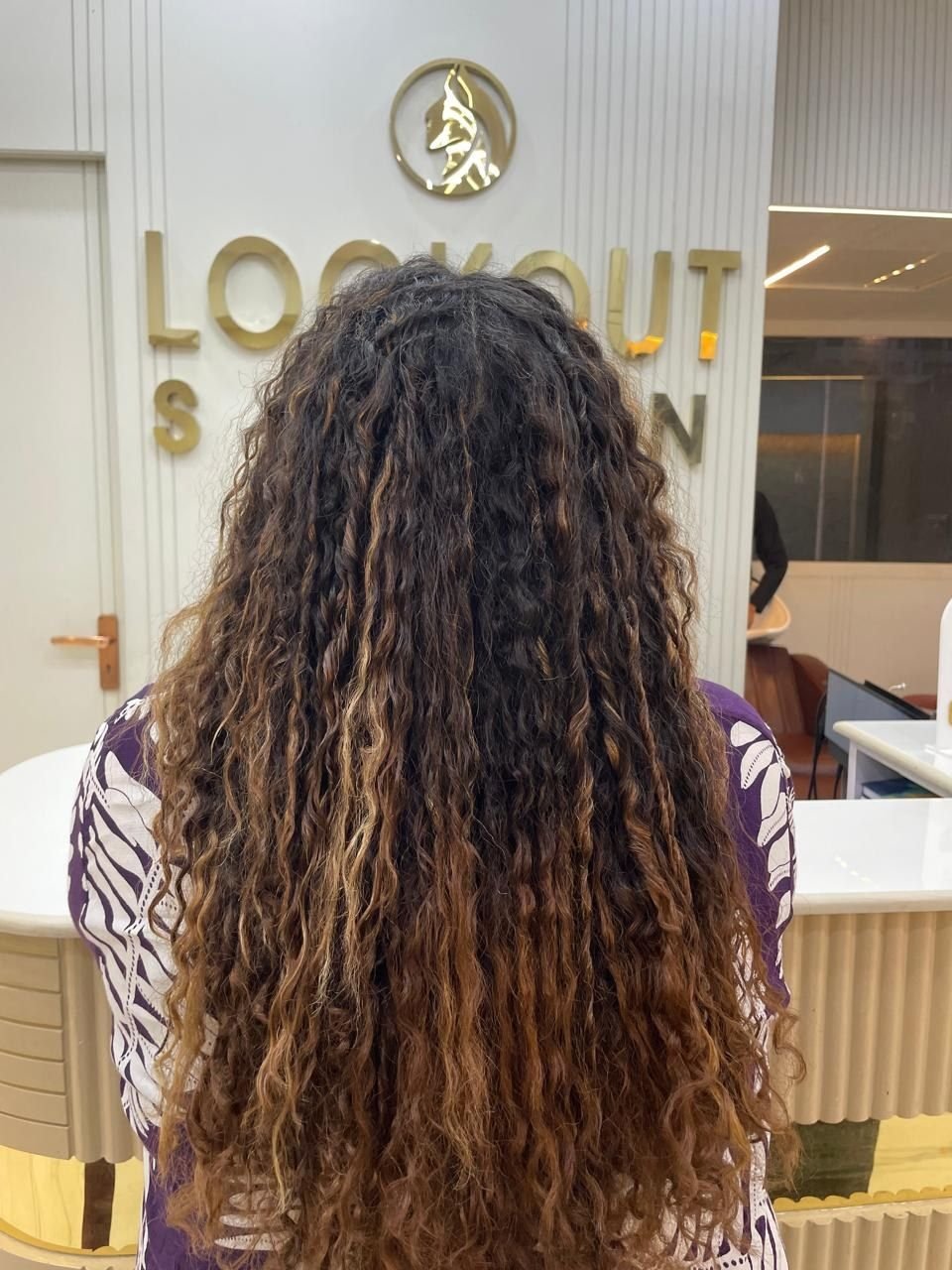 Back view of long curly hair with caramel highlights, in front of the LookOut Salon sign