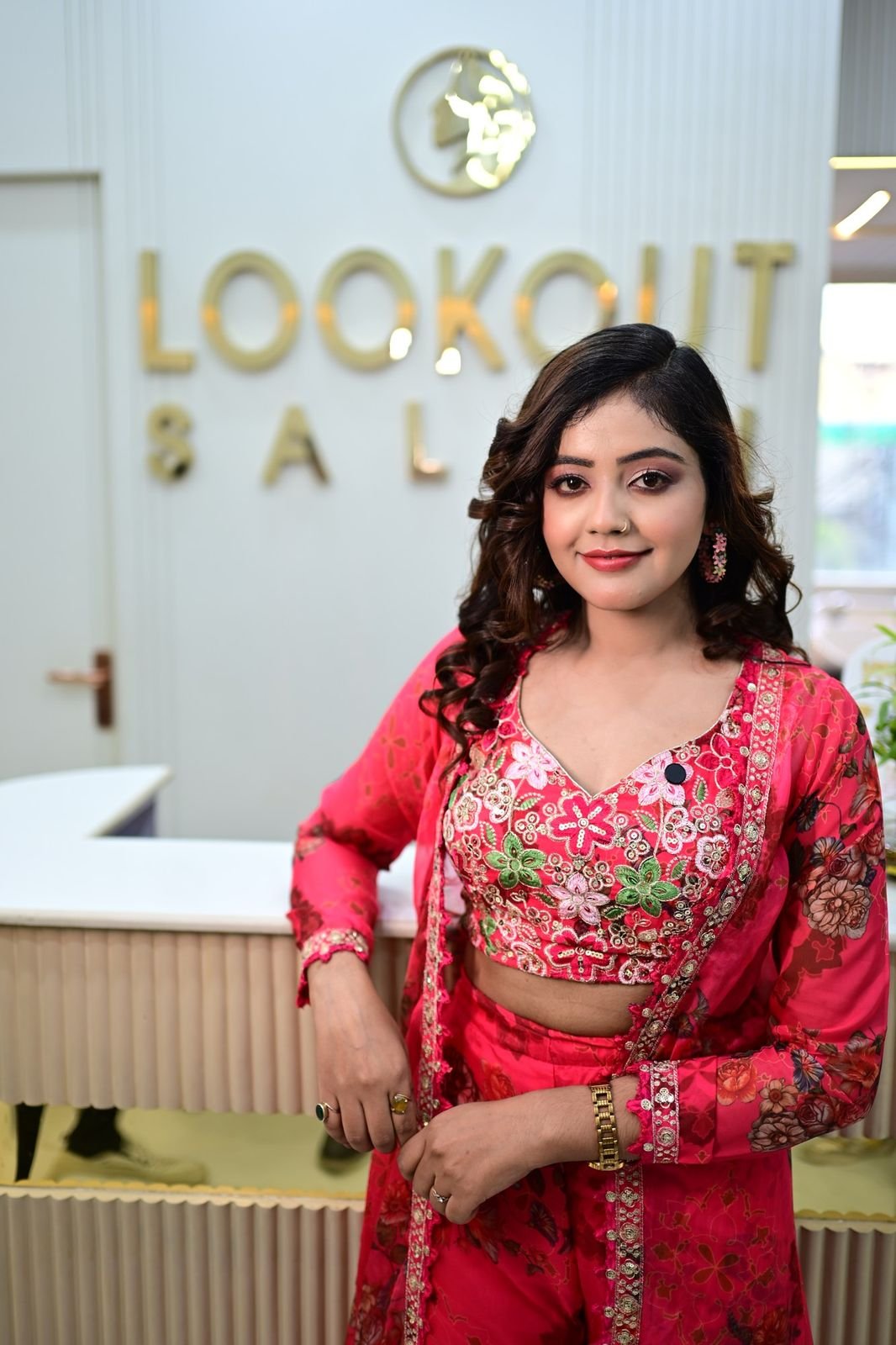 Woman with soft curls in a red and pink embroidered outfit, standing at the LookOut Salon reception